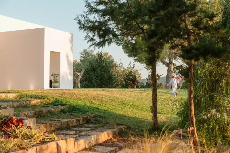 Modern white building on the left, surrounded by a serene garden. A child with a red toy airplane runs on the grassy slope, evoking joy and freedom.