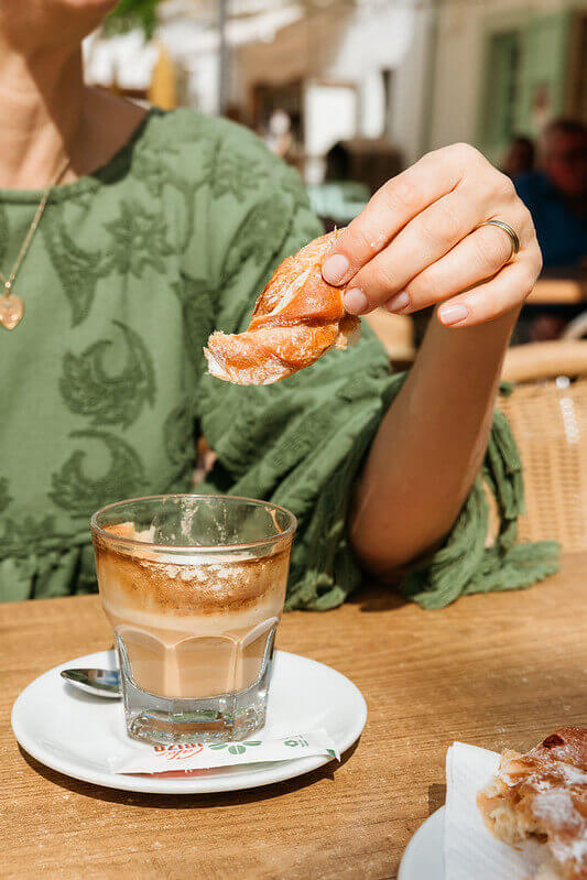 Woman holding a pastry beside a coffee at an outdoor cafe