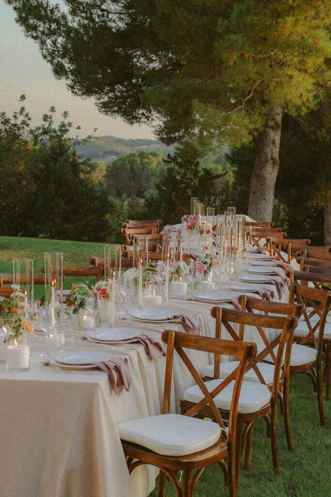 A long, elegantly set outdoor dining table with white tablecloths, cross-back wooden chairs, tall glass candle holders, and pink floral arrangements on a lawn at a luxury villa in Mallorca.