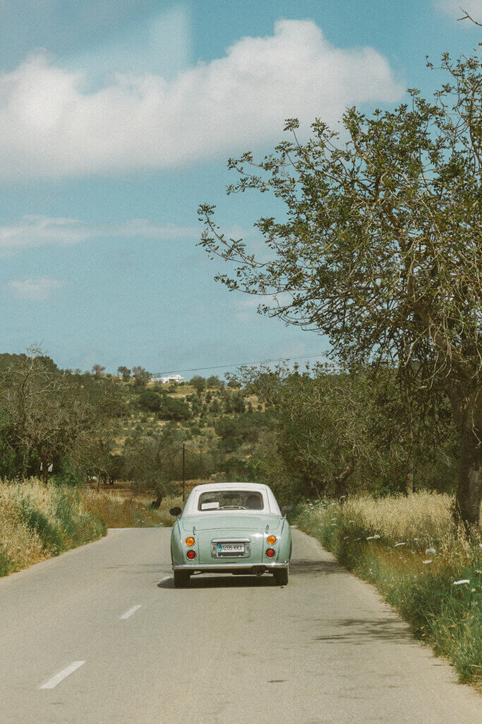A mint green vintage Nissan Figaro driving down a rural road toward a luxury villa on a hillside in Mallorca, Spain.