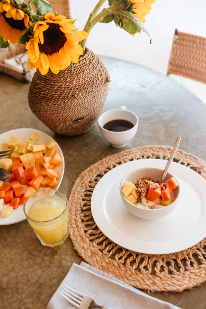 A tropical breakfast setup with a papaya fruit bowl, coffee, juice, and sunflowers on a rustic table in Mexico