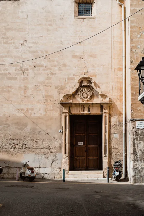 A weathered stone building with an ornate wooden door in a narrow street. Two scooters are parked nearby, casting long shadows. The scene feels historic and serene.
