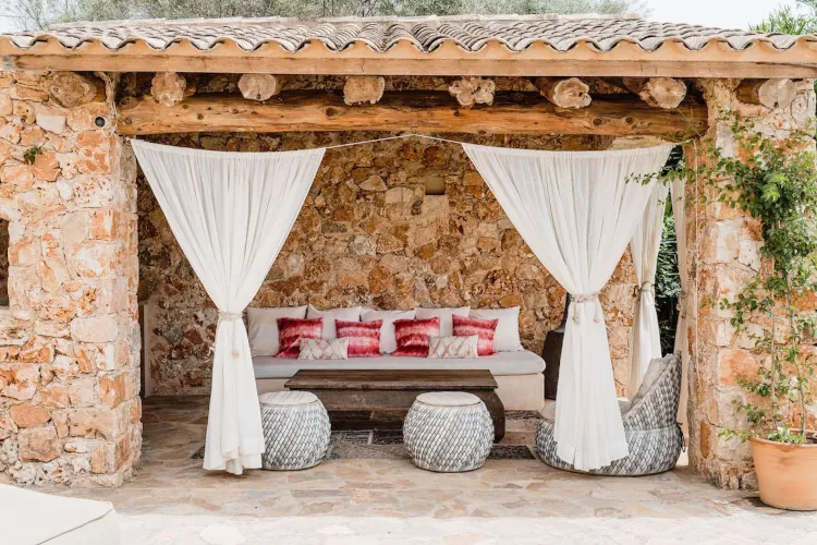 Stone cabana with rustic wooden beams and white drapes framing a cozy seating area. Cushions in pink hues adorn the bench. A serene, inviting vibe.