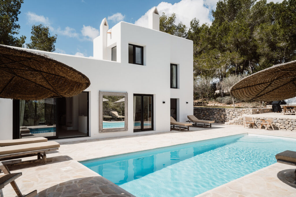 The pool terrace faces the white villa, with loungers and thatched parasols at Can Romero Villa in Santa Eulalia, Ibiza.