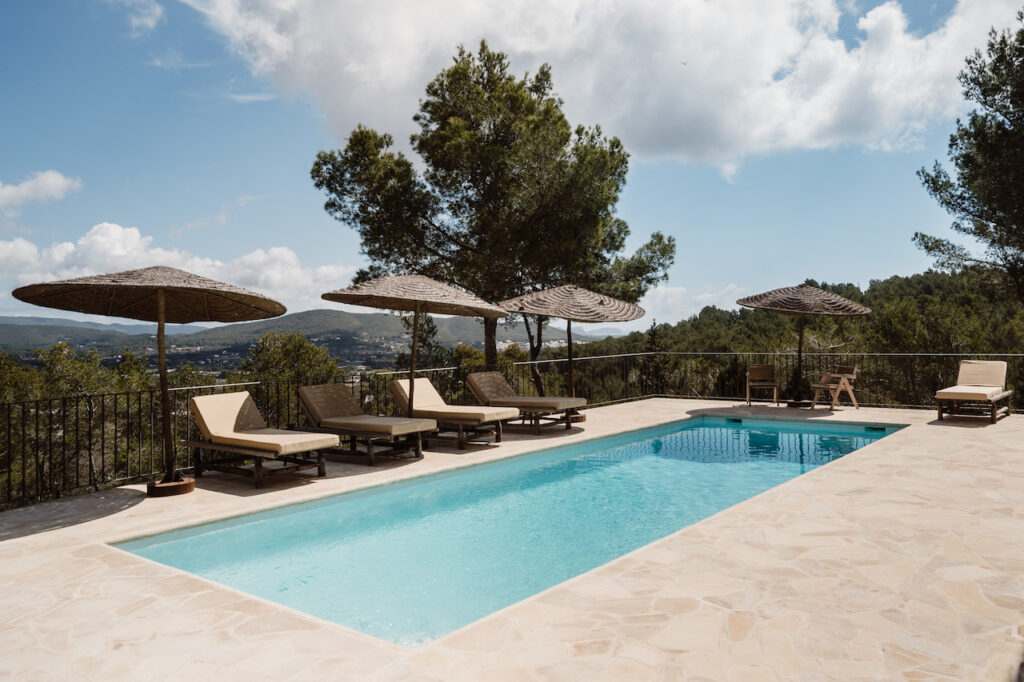 A turquoise pool and thatched parasols overlook wooded hills at Can Romero Villa in Santa Eulalia, Ibiza.