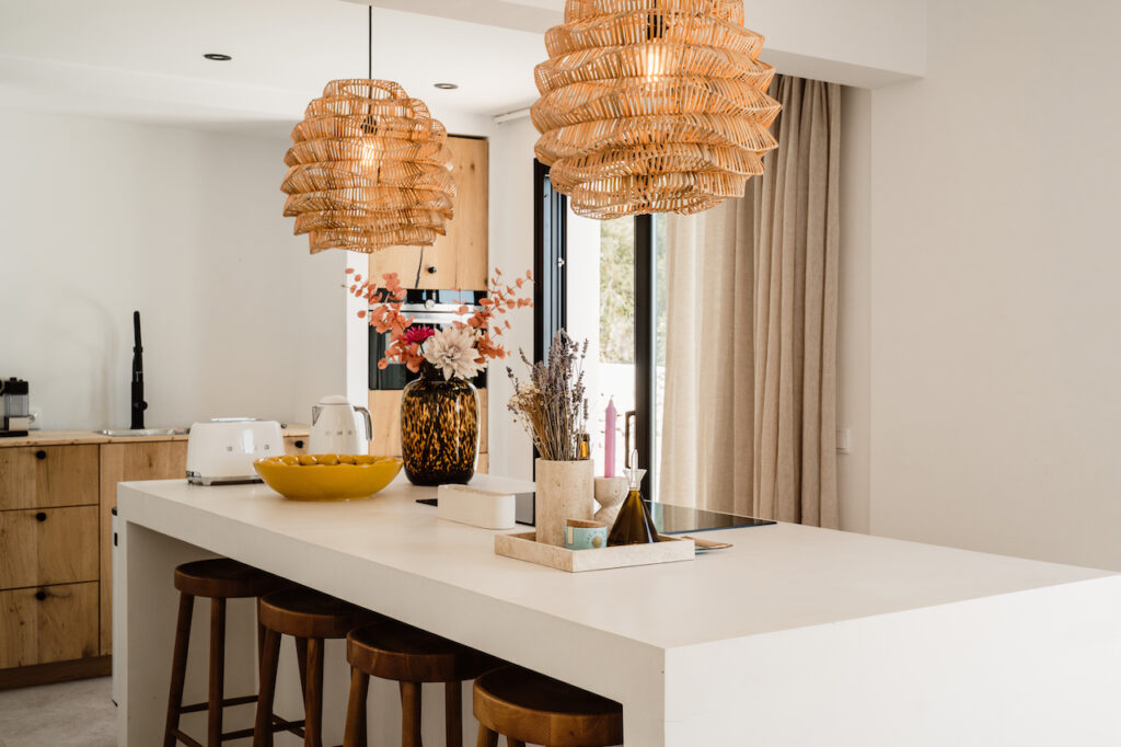 A white kitchen island with wooden stools sits beneath woven pendant lights at Can Romero Villa in Santa Eulalia, Ibiza.