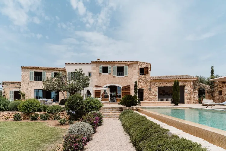Rustic stone villa with green shutters, surrounded by lush gardens. A clear blue sky overhead and a sparkling pool create a peaceful, inviting scene.
