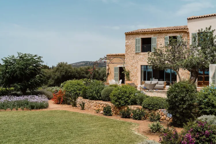 Rustic stone villa with green shutters, surrounded by lush gardens and lavender. Patio with cozy furniture. Calm, sunny atmosphere.