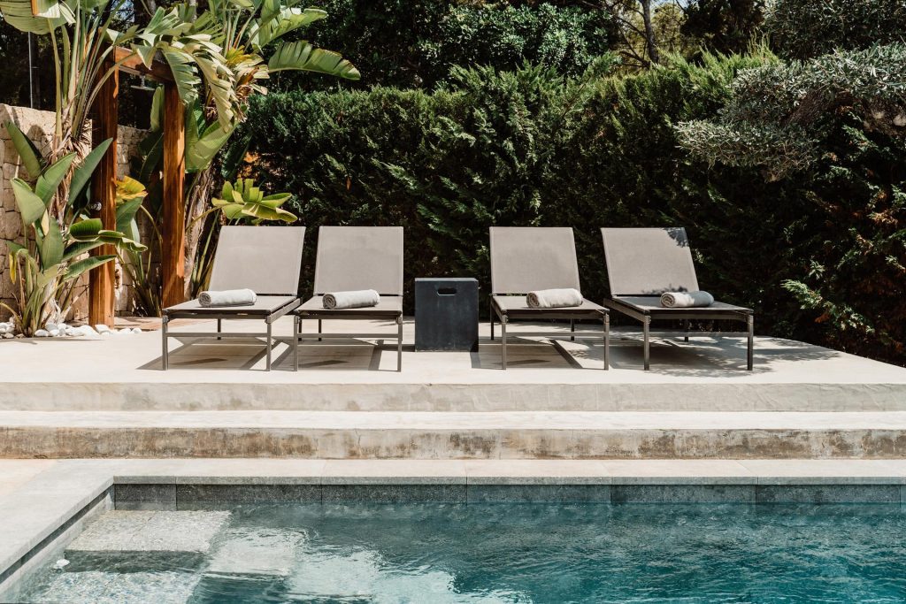Pool at Sa Brisa Villa in Santa Eulalia, Ibiza, with four sun loungers on a raised deck surrounded by greenery.