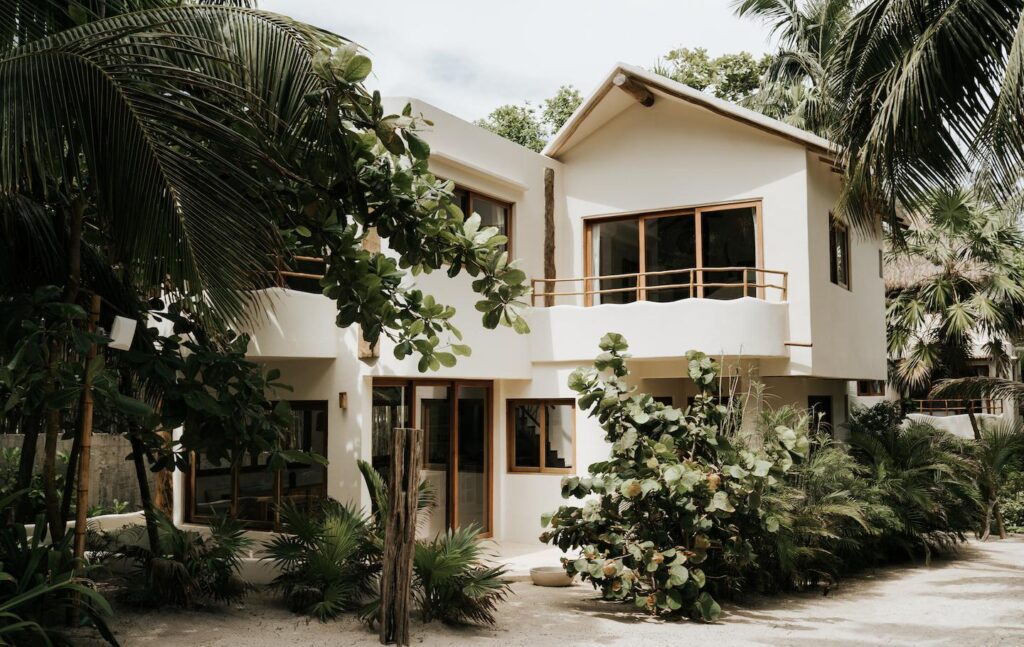 Wide view of a white villa with wood-framed windows and a small upper balcony, surrounded by tropical plants.