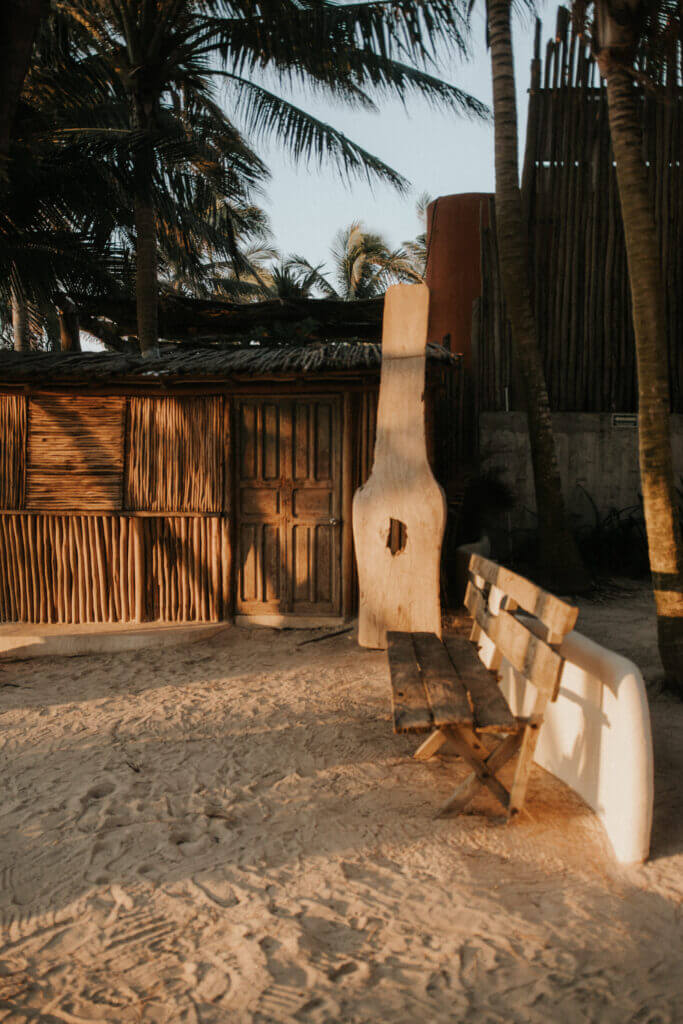 Sunlit sandy courtyard with a wooden bench beside a curved white structure, palm trees, and a rustic timber building.