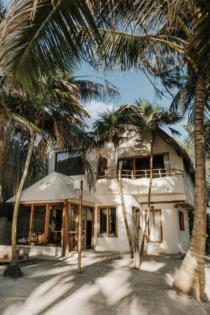 Two-story white villa with wood-framed windows and a thatched roof among palm trees.
