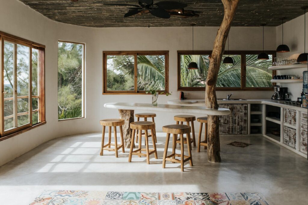 Bright kitchen with a curved counter, wooden stools, exposed tree-trunk supports, and windows overlooking palms.