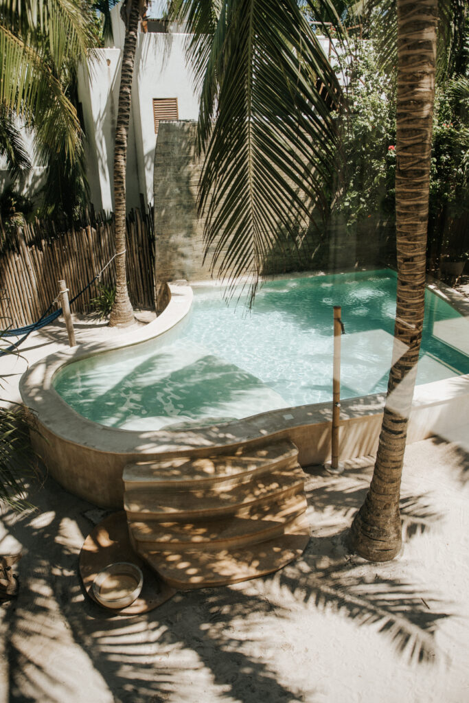 Curved turquoise pool shaded by palm trees, with rounded entry steps and a bamboo fence.
