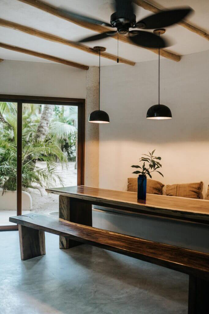 Long wooden dining table and bench beneath pendant lights, beside a garden-facing glass door.