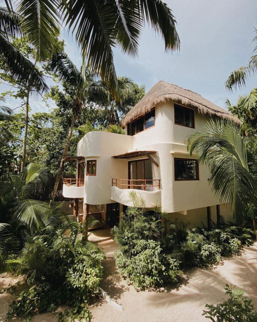 White villa with a thatched roof and curved balconies surrounded by palm trees.