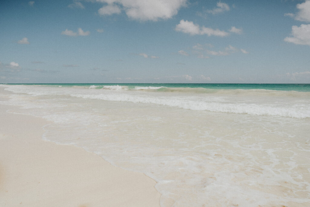 Gentle waves roll onto a wide, pale sand beach beneath a partly cloudy sky.