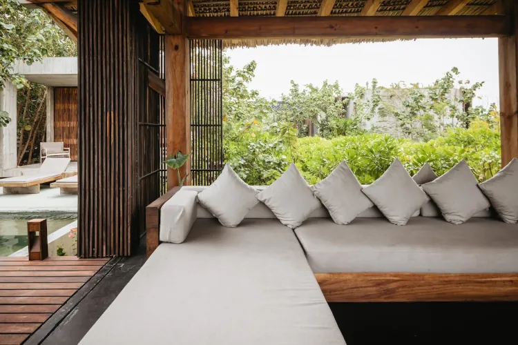 A view from inside a wooden, thatched-roof pavilion looking out toward a small pool. A long, grey L-shaped outdoor sofa with several white accent pillows is built into the wooden frame, providing a shaded view of the surrounding garden.