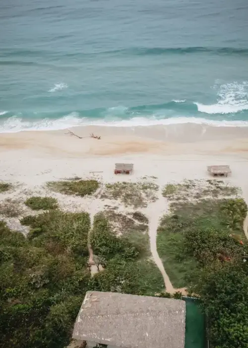 An aerial, high-angle view of a serene beach in Puerto Escondido, Mexico. The turquoise ocean waves crash onto light tan sand. Two small, thatched-roof palapas sit on the shoreline, with a narrow dirt path winding through green coastal vegetation toward a larger thatched building in the foreground.