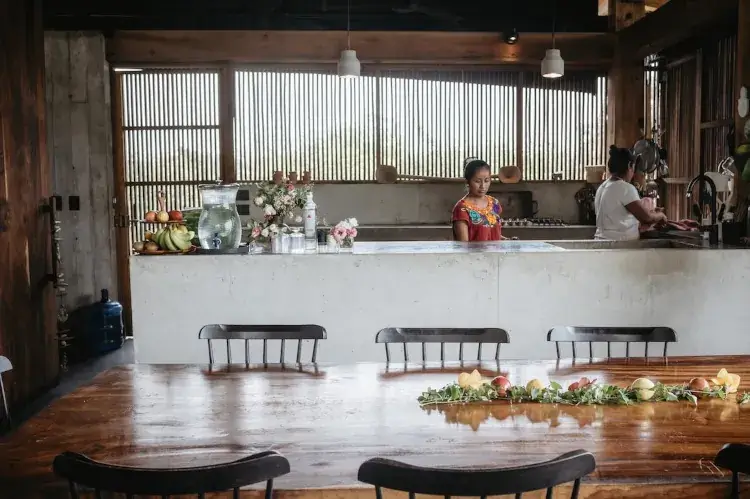 An open-concept luxury kitchen and dining area featuring a long wooden table and a minimalist concrete island. Two women are working in the background. The space is characterized by warm wood accents, slatted windows for natural light, and a collection of fresh fruit and flowers.