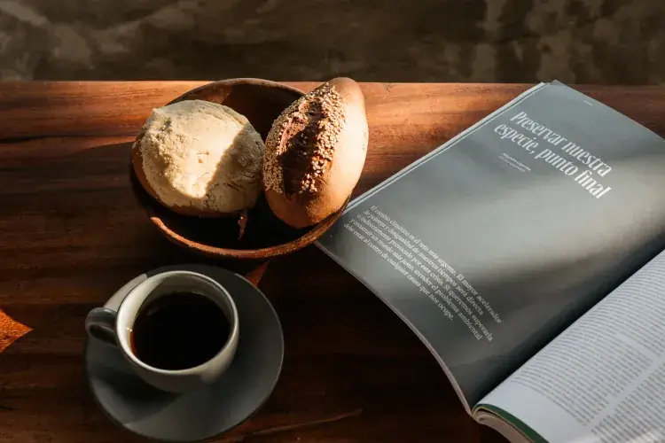 A top-down lifestyle shot of a morning setting on a wooden table. It features a cup of black coffee, a bowl of Mexican sweet bread (Concha and a bolillo), and an open magazine with a Spanish headline, all bathed in soft, natural sunlight.