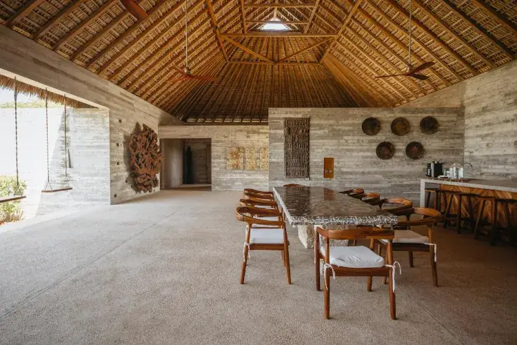 An expansive open-air room with a high thatched ceiling and exposed wood rafters features a long natural wood dining table surrounded by matching chairs with white cushions. A textured stone and concrete bar with barstools is to the right, and a hanging swing is visible to the far left. The concrete walls are decorated with large carved wood sculptures and a cluster of circular plates.