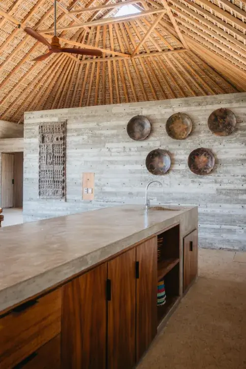 A close-up view focusing on a modern poured-concrete island with an integrated sink. Behind the island are wood-fronted lower cabinets and a light grey board-formed concrete wall featuring circular, weathered metal plates and a tall, intricate wooden carved panel. The high thatched ceiling with its geometric wood structure is clearly visible.