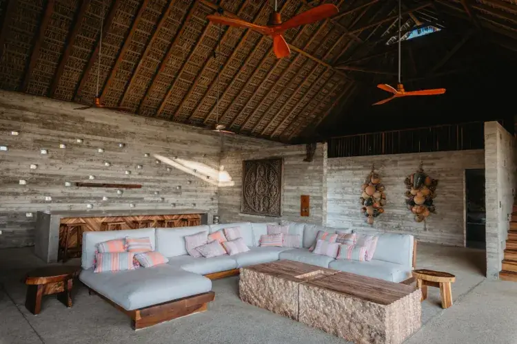A view into a large, comfortable open lounge with a soaring thatched ceiling and visible wooden rafters. A massive L-shaped sectional sofa in light grey with striped orange and purple cushions anchors the space, facing a large, rough-hewn rectangular rock and wood coffee table. To the left is a long, textured concrete bar with wooden stools, backed by a unique wall dotted with integrated metal lights and a carved wooden plaque. To the right, decorative bundles of rustic ceramic jars are mounted on the concrete wall next to a wooden staircase. The floor is textured concrete.