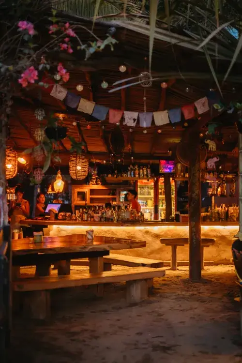 An atmospheric beach bar at night with a sandy floor and warm, glowing lanterns. Colorful prayer flags hang from the thatched ceiling over a wooden bar counter where a bartender stands ready to serve.