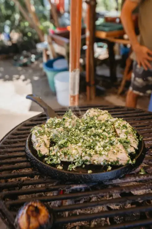 A close-up of fresh fish fillets heavily seasoned with minced garlic and herbs, cooking in a cast-iron skillet over an open-fire charcoal grill.