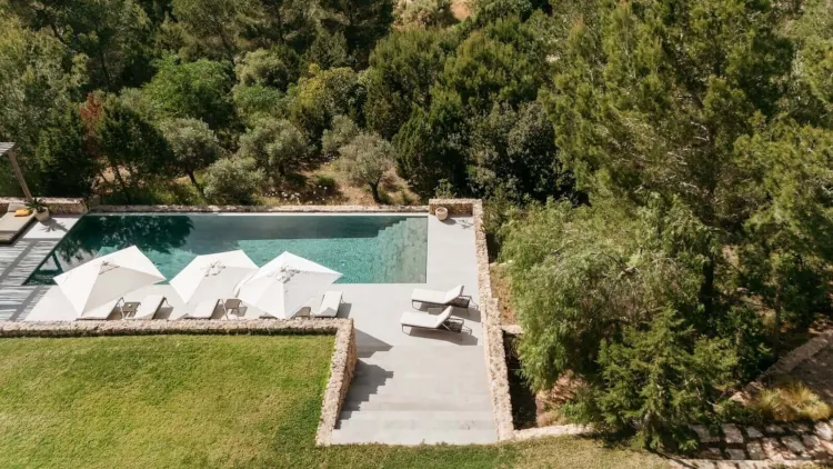 Aerial view of a luxurious pool with three white umbrellas, surrounded by lush greenery. Two lounge chairs are on the stone deck, creating a serene oasis.
