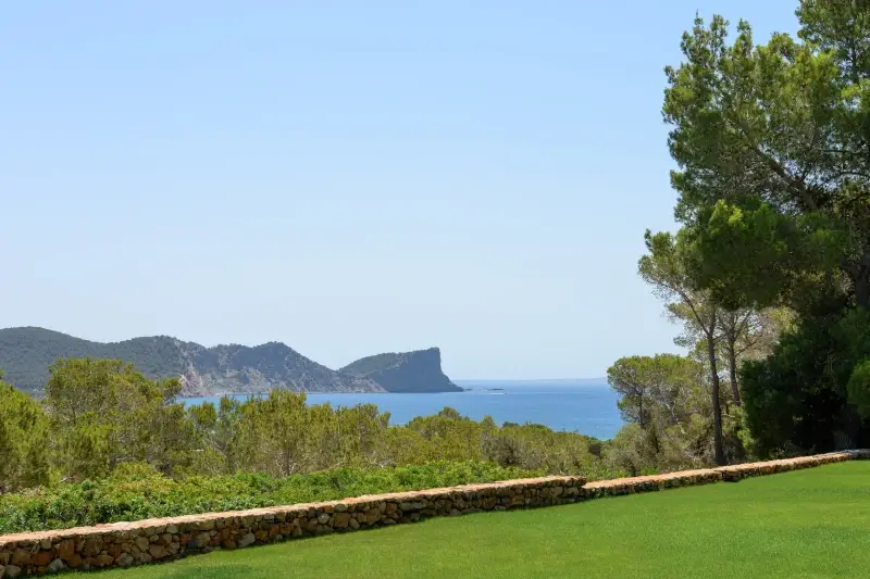 Scenic view of a grassy lawn bordered by a stone wall, leading to dense green trees. Beyond, a calm blue sea and distant rocky cliffs under a clear sky.
