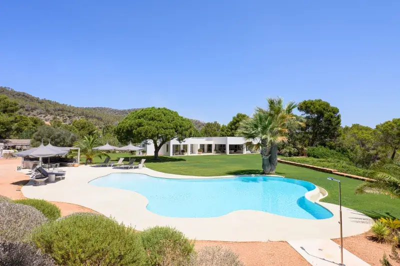 Luxurious villa with a modern design, surrounded by lush greenery. A large, clear blue pool with sun loungers and umbrellas creates a serene atmosphere.