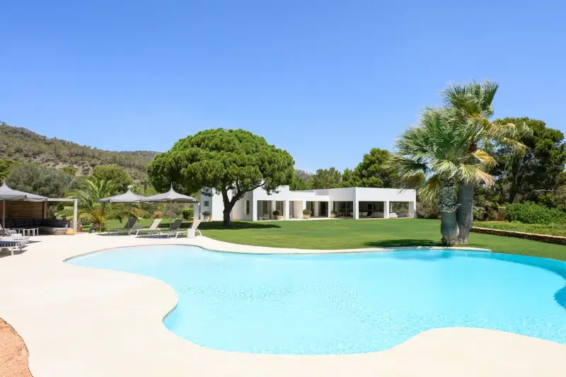 A luxurious, modern white villa is surrounded by lush green trees. In the foreground, a pristine, curved pool reflects the clear blue sky, evoking tranquility.