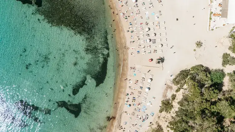 Aerial view of a sandy beach with turquoise water on the left and rows of colorful umbrellas and sunbeds on the right. Sparse greenery borders the beach area.