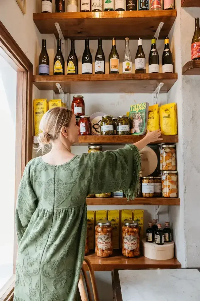 A woman in a green dress, viewed from behind, reaches for a yellow bag on wooden shelves filled with wine bottles, canned goods, and preserves. The setting appears cozy and inviting.