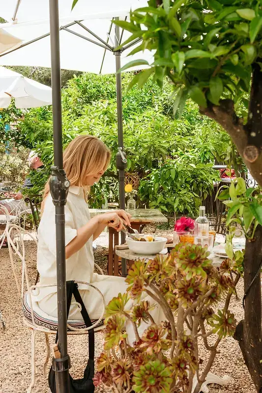 A woman in white sits at an outdoor cafe under umbrellas, surrounded by lush greenery. She's dining peacefully, conveying a serene and relaxed atmosphere.