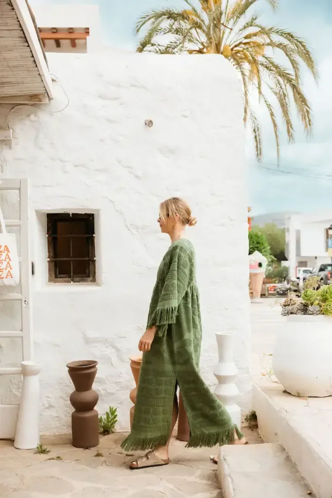 A woman in a green fringed dress walks past a white stucco building with planters. A palm tree is visible, suggesting a sunny, relaxed Mediterranean scene.