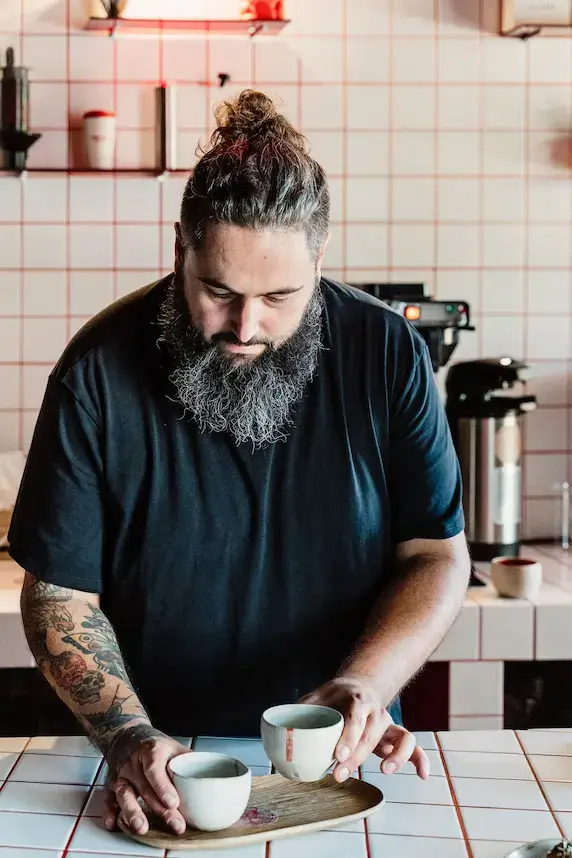 Barista with a beard and tattoos, wearing a black shirt, arranges cups on a tray in a coffee shop with white tiled walls. The scene is calm and focused.