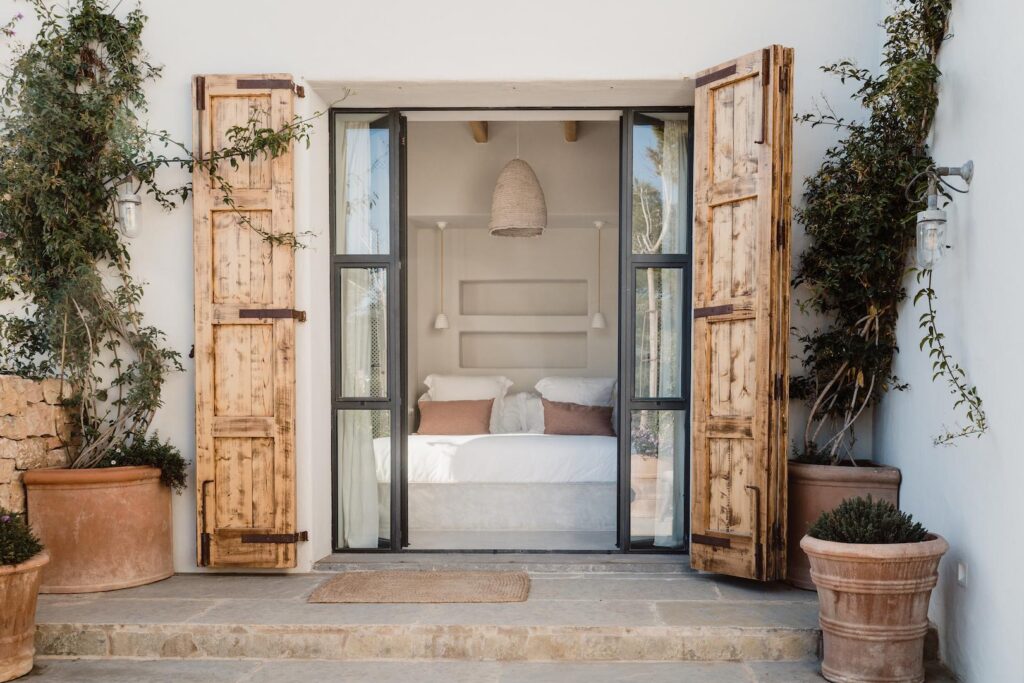 Open wooden shutters frame a white bedroom at Can Flores Villa in Cap Martinet, Ibiza.