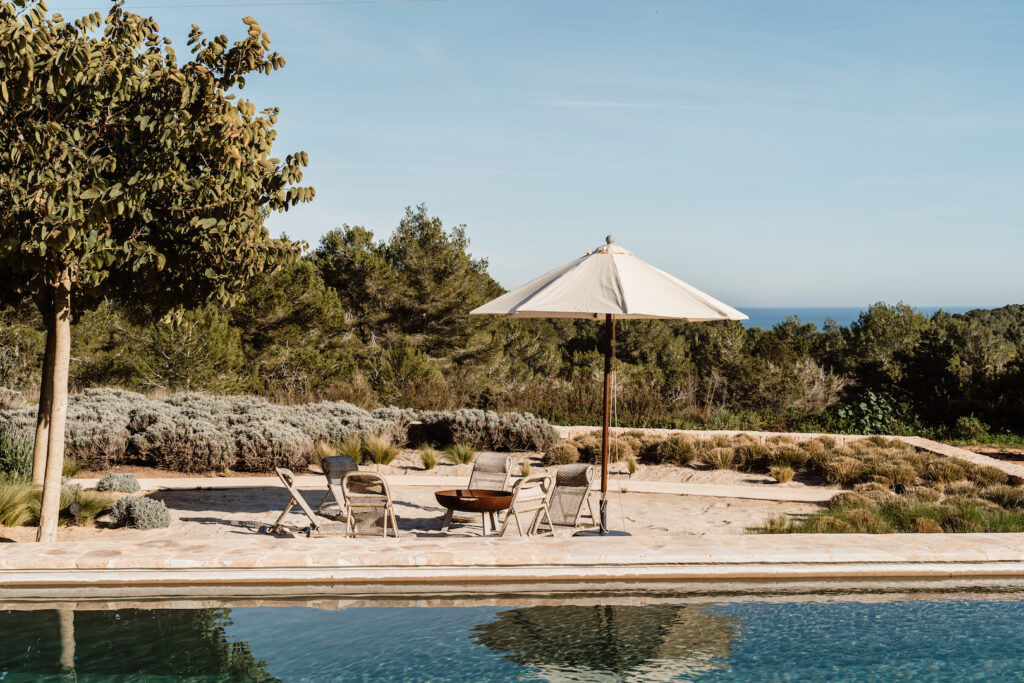 Poolside seating beneath an umbrella, with trees and a sea view at Can Flores Villa in Cap Martinet, Ibiza.