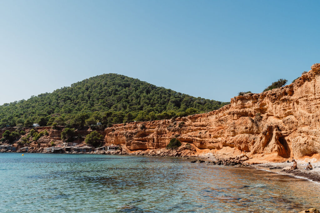 Sa Caleta Villa, Ibiza: calm water and reddish cliffs along a cove backed by a green hillside.