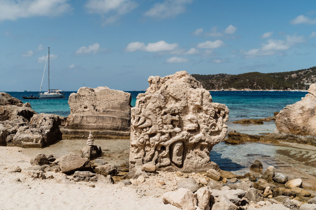 Sa Caleta Villa, Ibiza: carved stone sculptures on a rocky beach, with clear turquoise water and a sailboat offshore.