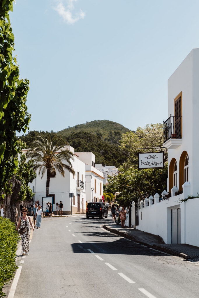 Sunny village street with white buildings, palm trees, pedestrians, and a café sign.