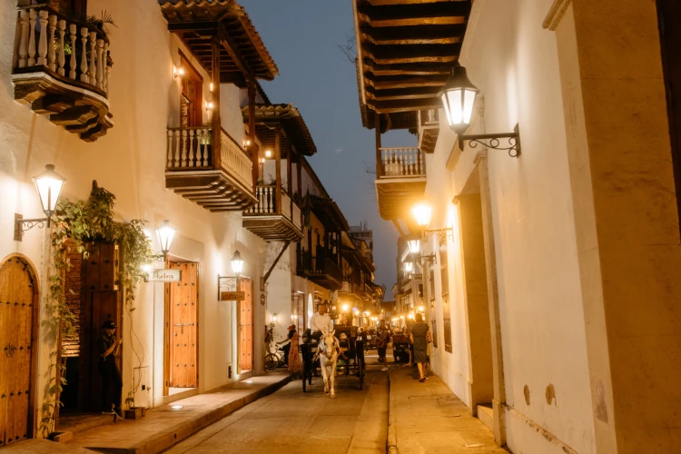 A charming cobblestone street at dusk in Cartagena, Colombia, lit by warm lanterns and lined with historic balconies. A horse-drawn carriage adds a nostalgic feel.