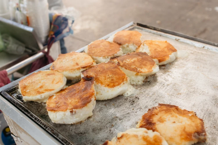 Golden, fluffy pancakes sizzle on a flat griddle, emitting warmth and savory aromas. The street vendor's setting adds an inviting, rustic feel.