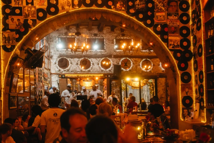 Cozy bar scene with warm, ambient lighting, featuring musicians playing on stage through an arched entrance. Walls adorned with vinyl records and photos.