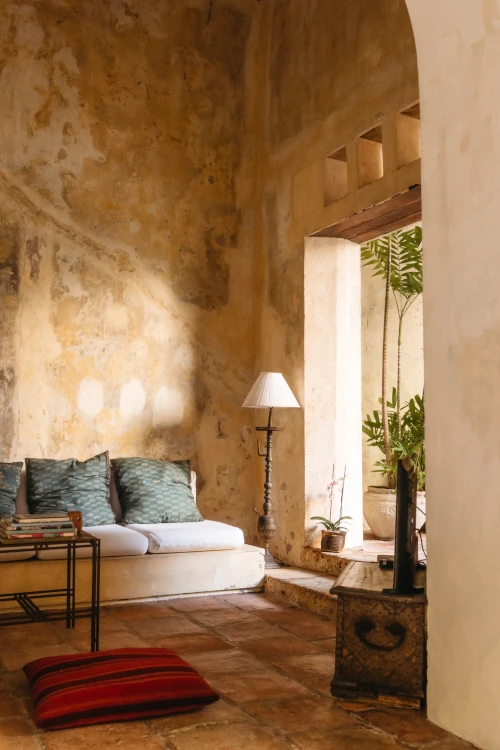 Cozy rustic room with textured beige walls and terracotta floor. Features a white sofa with blue cushions, a floor lamp, a potted plant, and a red striped cushion. Warm, relaxed ambiance.