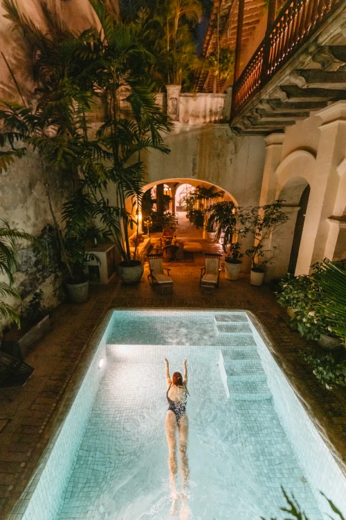 A person swims in a narrow, illuminated pool at night, surrounded by lush green plants and rustic stone walls. Warm, inviting light glows from an archway.