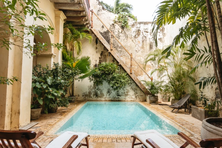 Cozy courtyard with a small, square pool surrounded by lush greenery. Worn stone walls and steps add rustic charm, evoking a serene, inviting ambiance.